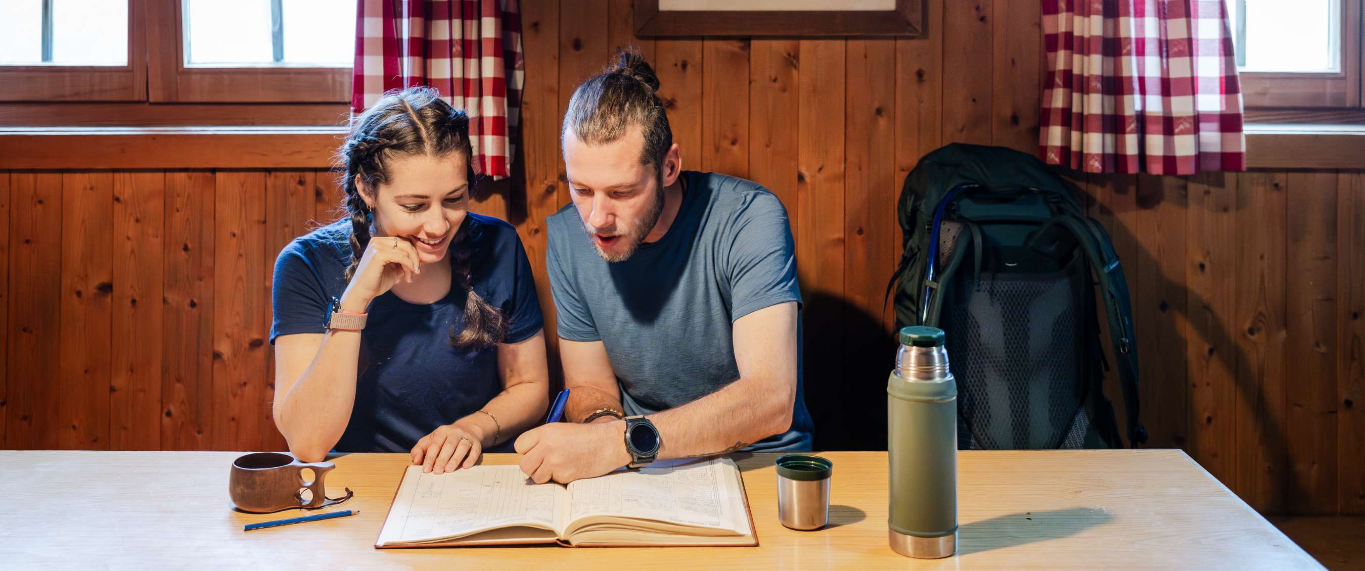 Man en vrouw bekijken een kaart in een berghut, met thermos en rugzak naast de tafel.